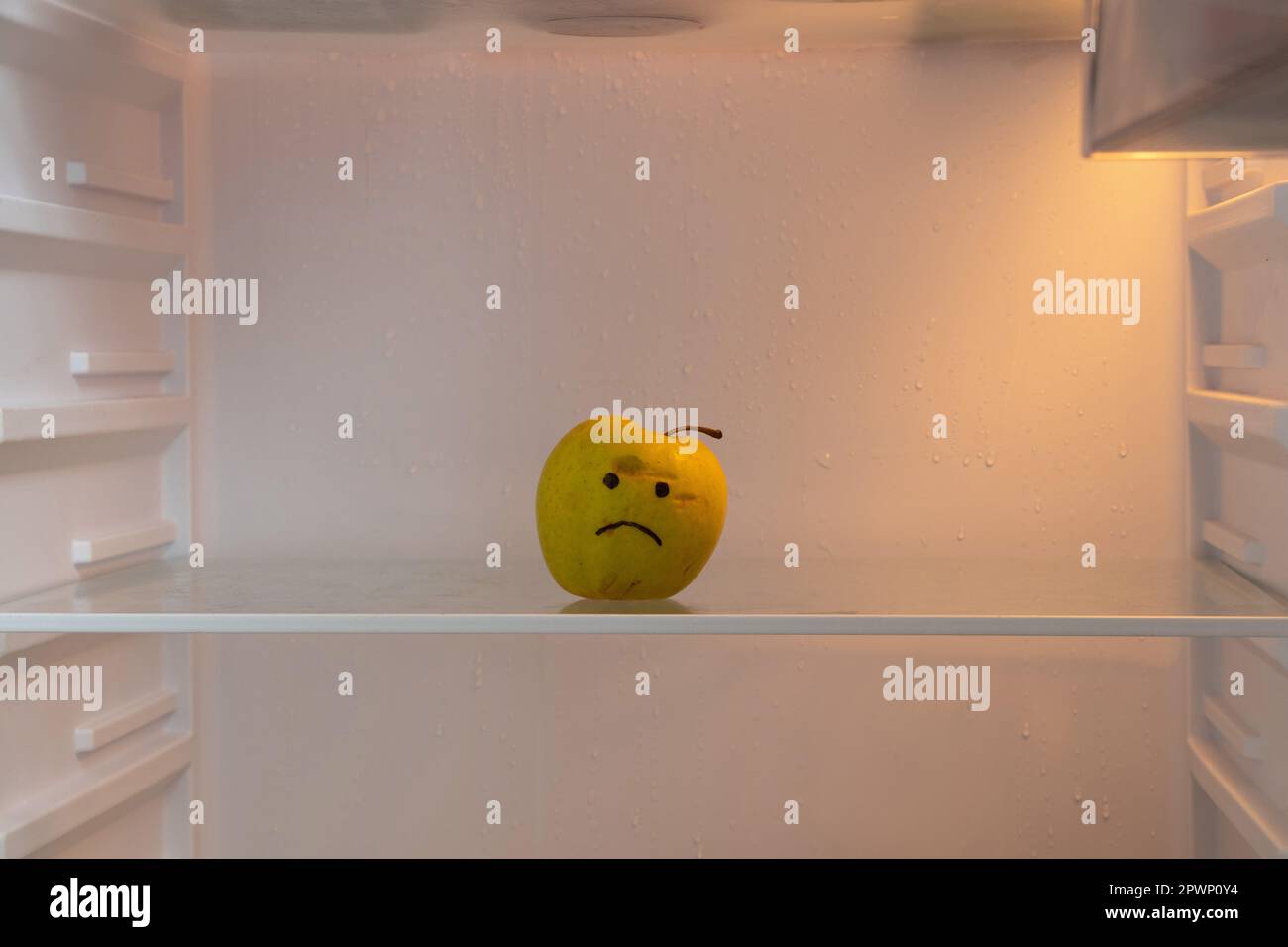 Ein trauriges Gesicht wird auf einen grünen Apfel gemalt, der im Kühlschrank steht, auf einem leeren Regal, Hunger und nichts zu essen, schlechte Laune Stockfoto