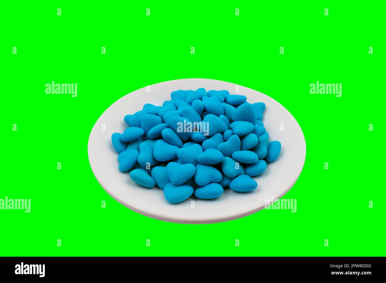 Weiße Platte mit blauen herzförmigen Süßigkeiten (Nahaufnahme) auf grünem Hintergrund (Chroma-Taste). Blick von oben Stockfoto