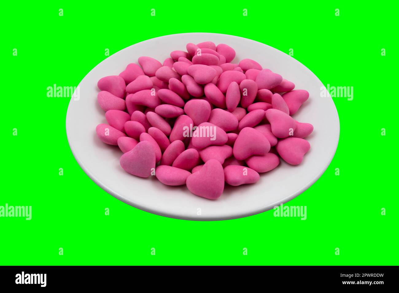 Weiße Platte mit rosafarbenen herzförmigen Süßigkeiten (Nahaufnahme) auf grünem Hintergrund (Chroma-Taste). Blick von oben Stockfoto