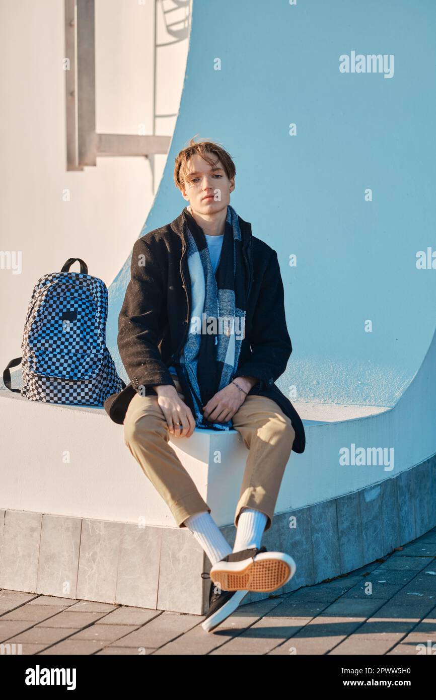 Junge Schülerin mit Rucksack in altem Wollmantel, Schal und kurzer Hose sitzt im Freien Stockfoto