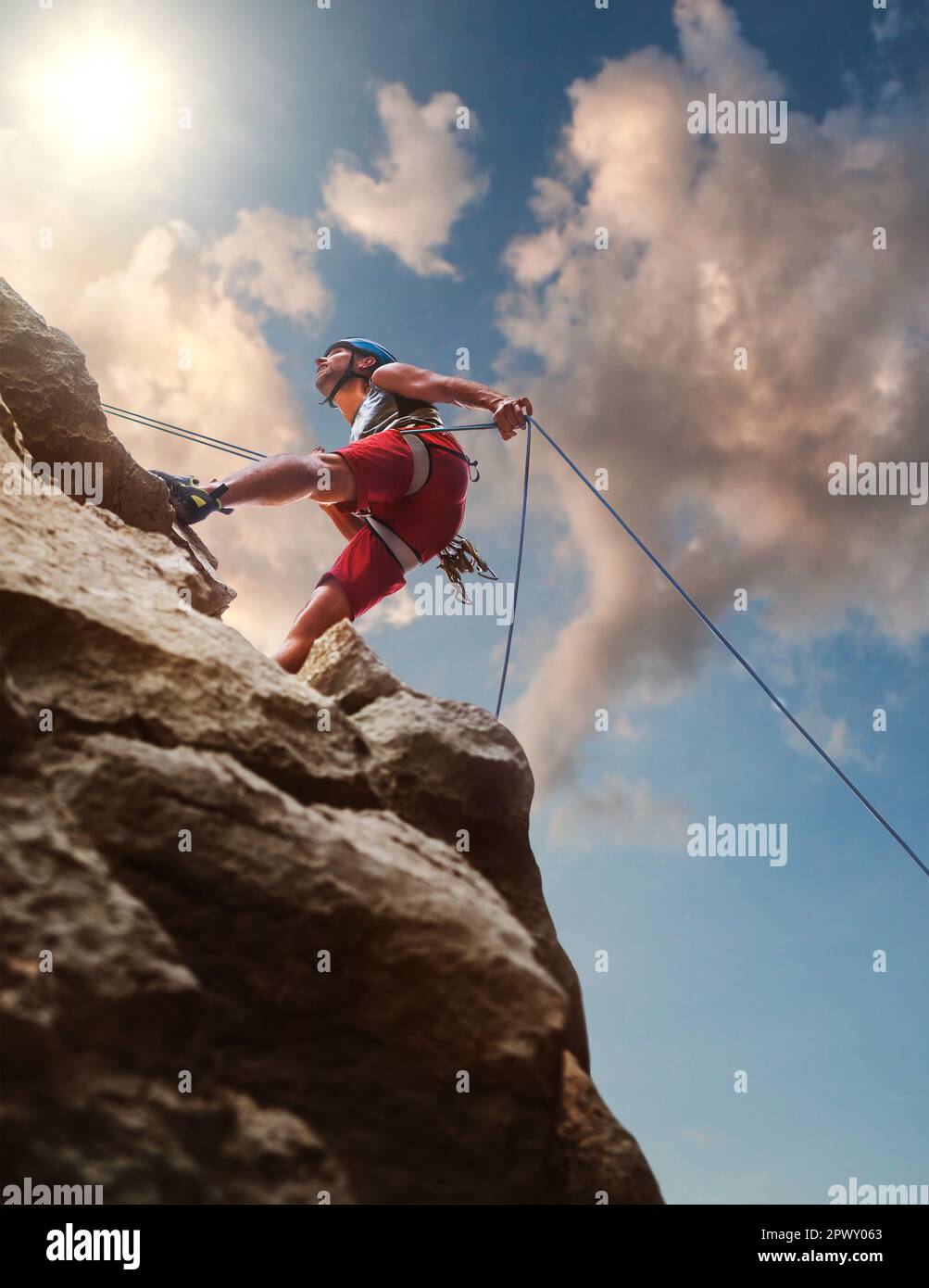 Muskulöser Kletterer Mann in Schutzhelm Abseilen von Felswand mit Seil Belay Gerät und Klettergurt auf Abend Sonnenuntergang Himmel Hintergrund Stockfoto