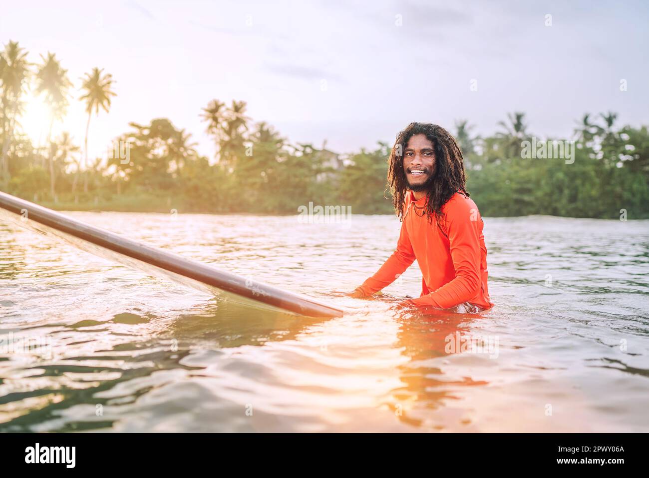 Schwarzer langhaariger Teenager-Mann, der auf einem langen Surfbrett schwimmt und auf eine Welle wartet, die zum Surfen bereit ist, mit Palmenhain-Sonnenstrahlen. Extreme Wassersportarten o Stockfoto