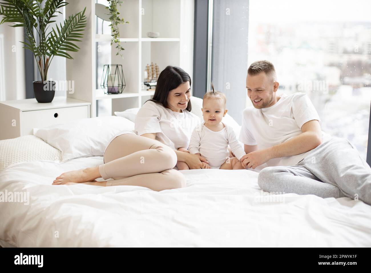 Kaukasische Eltern, die mit dem Baby auf dem Bett liegen und sich von beiden Seiten zärtlich umarmen. Süßes kleines Mädchen mit lustigem Pferdeschwanz, das leicht lächelt und nach vorne schaut. Konzept von Liebe und Familienschätzen. Stockfoto