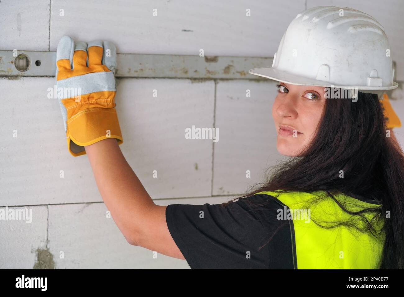 Junge brünette Bauarbeiterin mit weißem Schutzhelm und grüner Warnweste, Messwand mit Wasserwaage, Blick zurück Stockfoto
