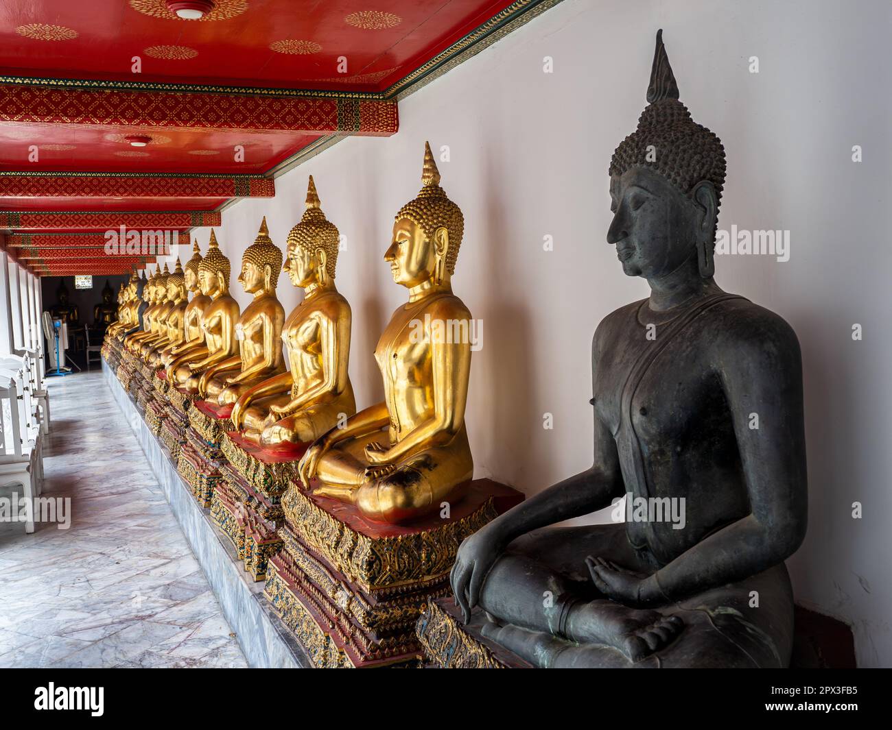 Goldene Buddha-Statuen in einer langen Schlange mit einer schwarzen Statue, die sich entlang einer Mauer am Wat Pho Tempel in Bangkok, Thailand, erhebt. Stockfoto