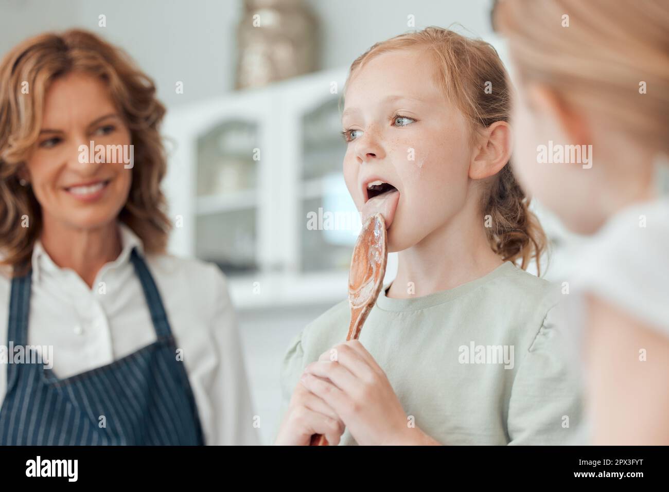 Ich muss probieren, um sicher zu gehen, dass es lecker ist. Ein kleines Mädchen, das einen Holzlöffel leckt, während es mit ihrer Schwester und Großmutter zu Hause backt Stockfoto