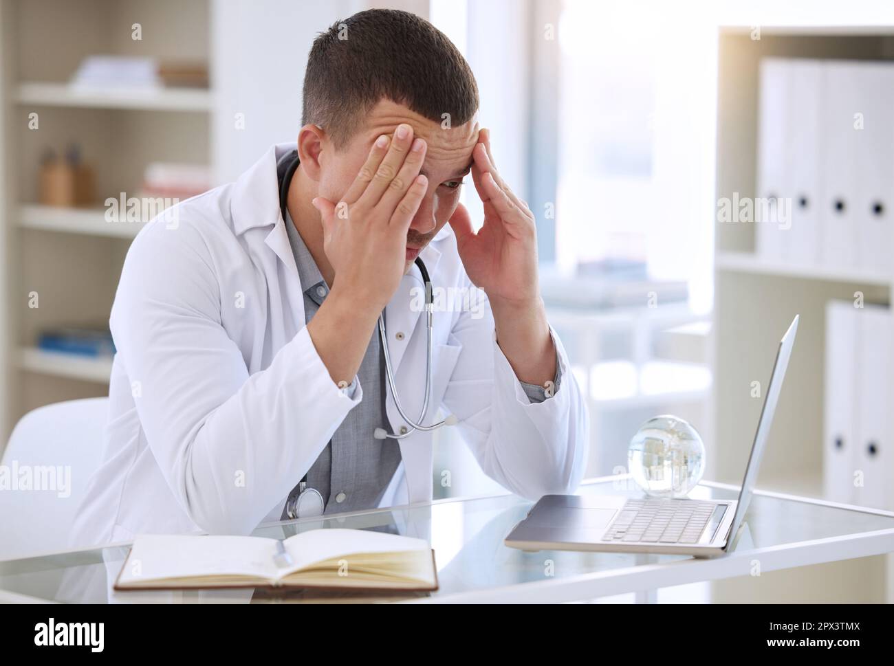 Ich kann bei diesen Kopfschmerzen nicht klar denken. Ein gutaussehender junger Arzt, der allein in seiner Klinik sitzt und sich gestresst fühlt, während er seinen Laptop benutzt Stockfoto