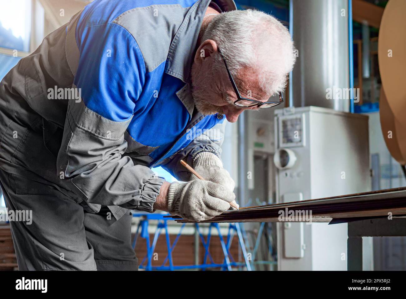Ältere Brillenarbeiter arbeiten im Geschäft. Ein Mann in Arbeitskleidung wird in der Werkstatt über die Werkbank gebogen und macht Markierungen oder Zeichnungen auf einem Metallblech. Industrieller Hintergrund. Stockfoto