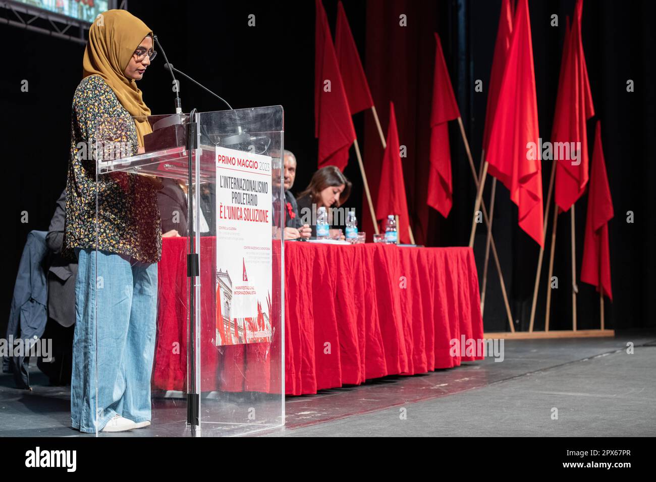 Eine Aktivistin von Lotta Comunista auf der Bühne während ihrer Rede mit den roten Fahnen im Hintergrund. Wanderarbeiter aus den römischen Vororten waren Ehrengäste bei der traditionellen Feier zum Mai, die Lotta Comunista, die exparlamentarische, internationalistische Bewegung, jedes Jahr im Teatro Brancaccio in Rom organisiert. Die wichtigsten Leitmotive der diesjährigen Konferenz waren ein entschiedenes Nein zum Frieden mit Waffen, die entschiedene Ablehnung jeglicher Form von Fremdenfeindlichkeit und die Notwendigkeit, die Proletarier der ganzen Welt zu vereinen. Stockfoto