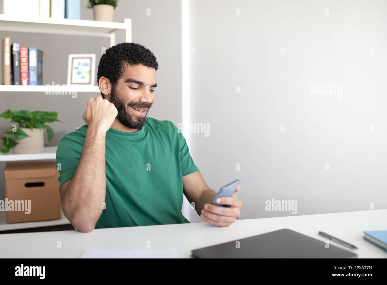Ein glücklicher junger Blogger aus dem Nahen Osten am Tisch mit Laptop schaut auf das Telefon und macht ein Siegeszeichen Stockfoto