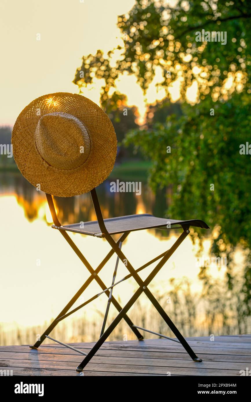 Ein Strohhut hängt auf einem Stuhl im Freien, steht auf einer Terrasse neben einem See bei Sonnenuntergang Stockfoto