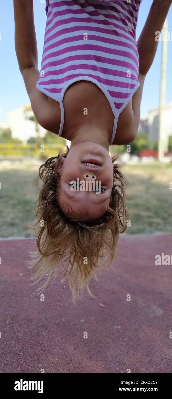 Das Mädchen steht auf dem Kopf. Ein Kind mit blonden Haaren in einem gestreiften rosa Kleid hängt an einer Bar auf dem Spielplatz. Die Haare hängen nach unten, der Mund ist halb offen. Gir Stockfoto