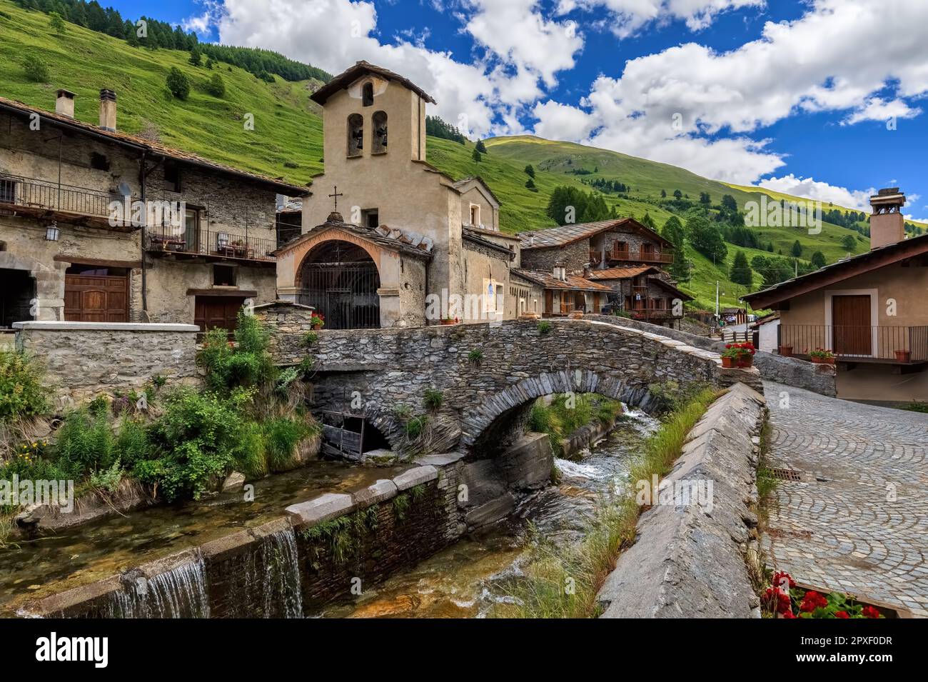 Blick auf die kleine Stadt mit alten Häusern, Pfarrkirche und mittelalterliche Brücke über den alpinen Bach als Berge im Hintergrund in Chianale, Italien. Stockfoto