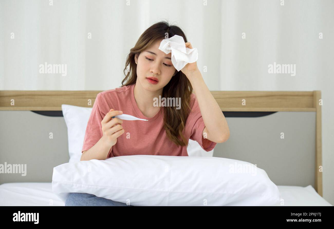 Junge asiatische Frau, die im Bett mit einem Digitalthermometer die Temperatur misst. Covid-19-infizierte bleiben allein in ihren Zimmern, nicht aus Sicherheitsgründen Stockfoto