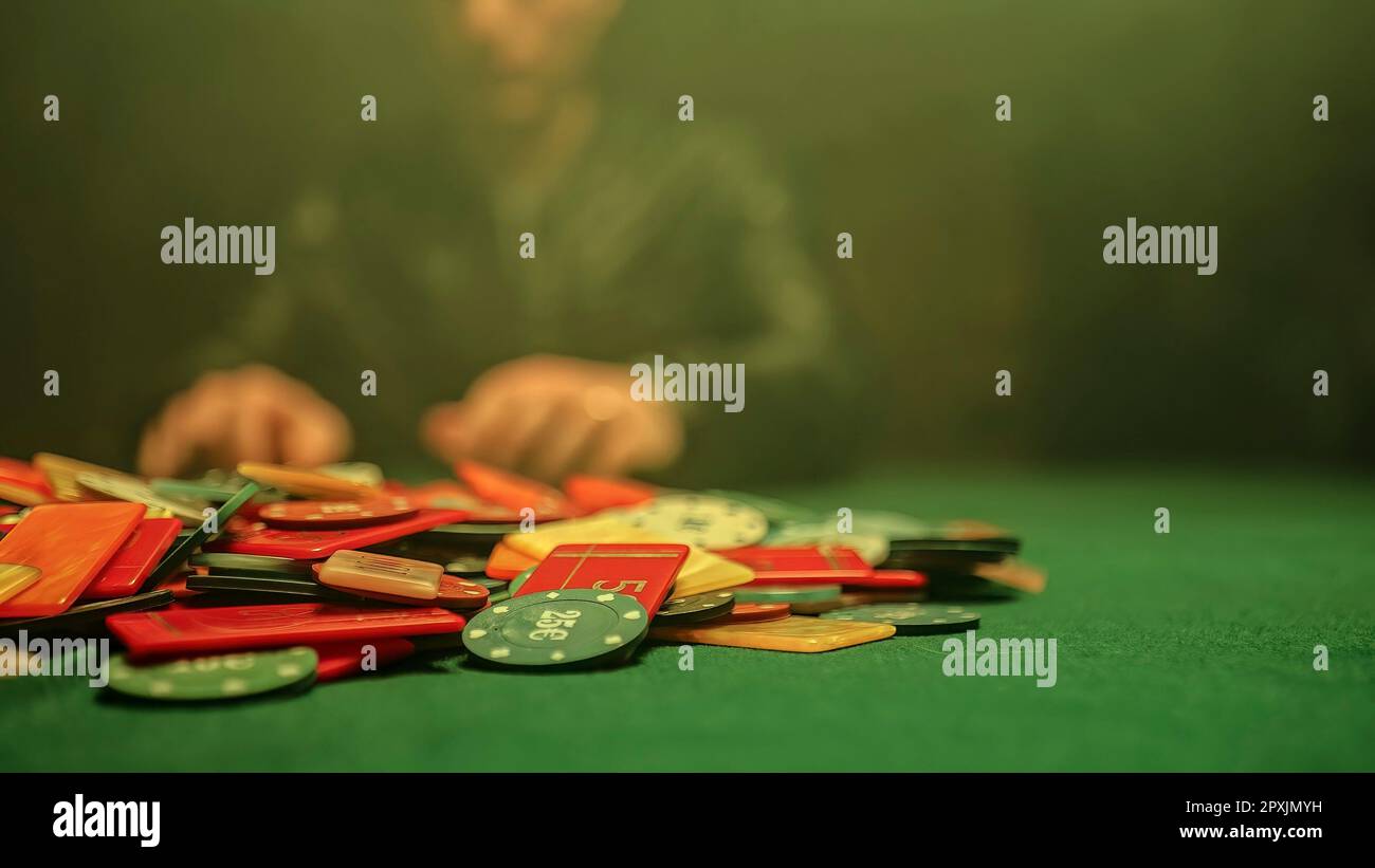 Eine chaotische Szene eines Pokerspiels mit Chips, die willkürlich über den Tisch verteilt sind. Verschwommener Hintergrund eines Pokerspielers, der am Tisch sitzt. Stockfoto