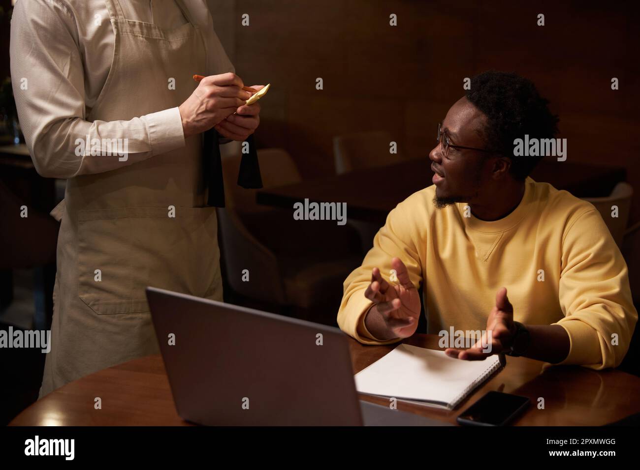 Ein junger afroamerikanischer Mann bespricht das Menü mit dem Kellner, während er sich während des Mittagessens im Café Notizen macht Stockfoto