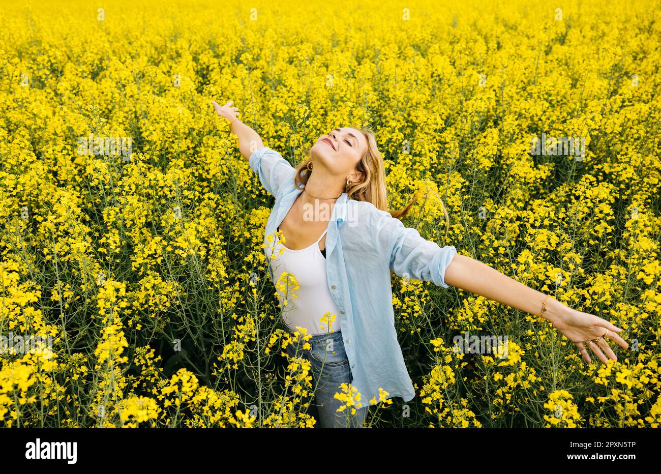 Wunderschöne, junge blonde, fröhliche Frau, mit ausgestreckten Armen, inmitten eines Feldes blühender gelber Rapsblumen Stockfoto