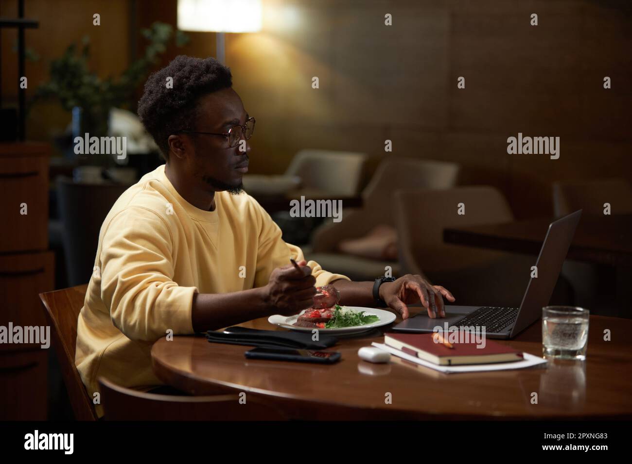 Afroamerikanischer Geschäftsmann, der online an einem Laptop arbeitet, am Tisch sitzt und im Café zu Mittag isst Stockfoto