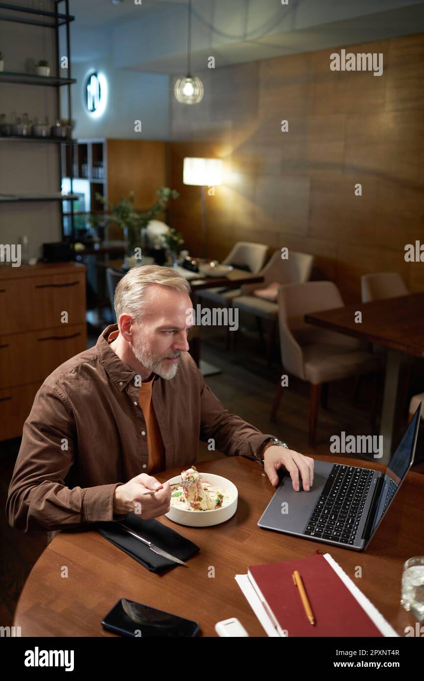 Ein erwachsener Mann, der während des Mittagessens ein Notebook benutzt und im Restaurant am Tisch sitzt Stockfoto