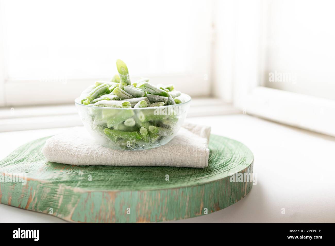 Tiefgefrorene grüne Bohnen in einer Glasschüssel auf grauer Serviette auf grünem Holzständer auf weißem Fenster in der Nähe des Fensters. Gefrorenes Gemüse. Gesunde Bio-Lebensmittel. Sust Stockfoto