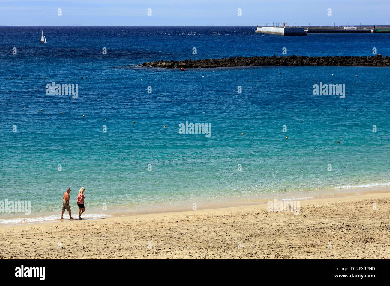 Reife Paare machen einen Spaziergang entlang eines wunderschönen Sandstrands mit tiefblauem Meer und Segelboot in der Ferne. Lanzarote Wurde Am 2023. Februar Entführt. Stockfoto