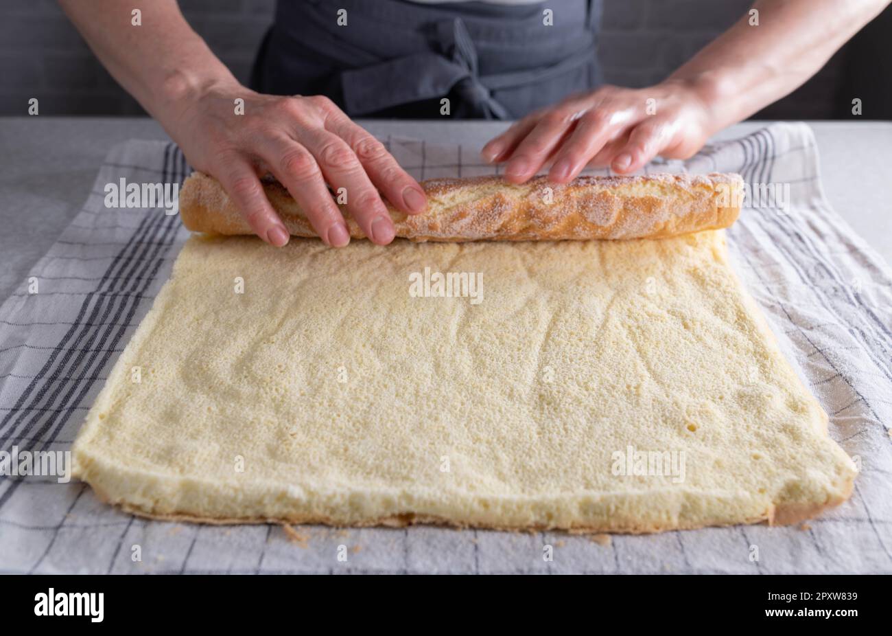 Frisch gebackener Biskuitkuchen für schweizer Brötchen, zum Abkühlen aufgerollt auf einem zuckerhaltigen Geschirrtuch von Frauenhänden Stockfoto