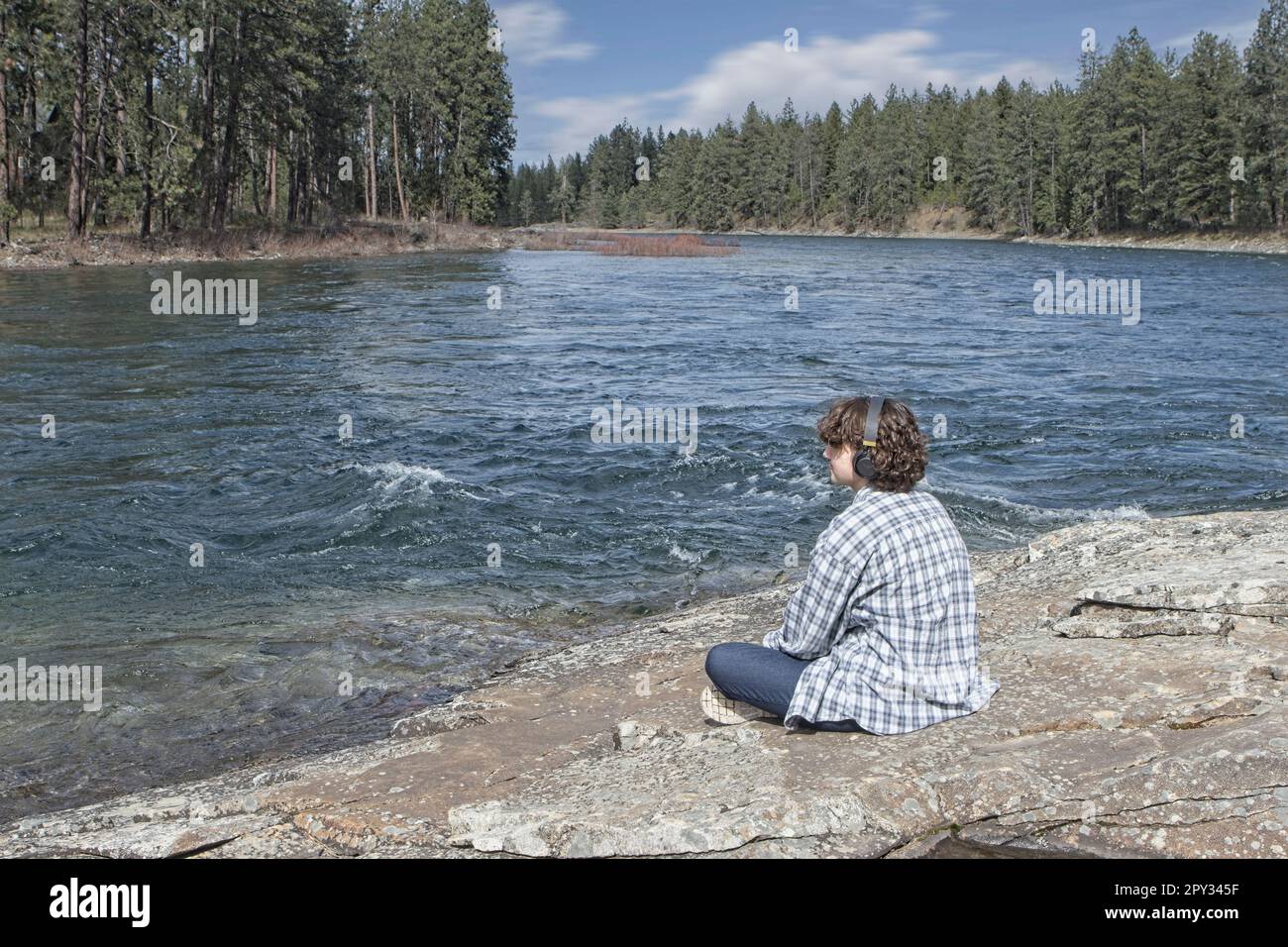 Eine junge Frau sitzt am Spokane River in Post Falls, Idaho, und entspannt sich an einem sonnigen Tag und hört Musik über ihre Kopfhörer. Stockfoto