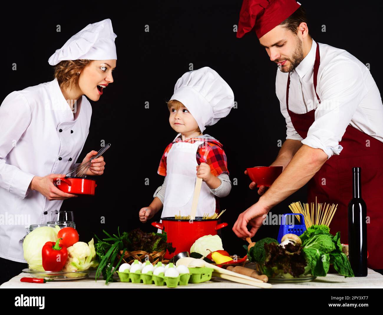 Glückliche Familie bereitet gesundes Essen zusammen in der Küche zu. Ein bezaubernder Sohn mit Kochmütze und Eltern, die zusammen zu Hause kochen. Mutter, Vater und Sohn Stockfoto
