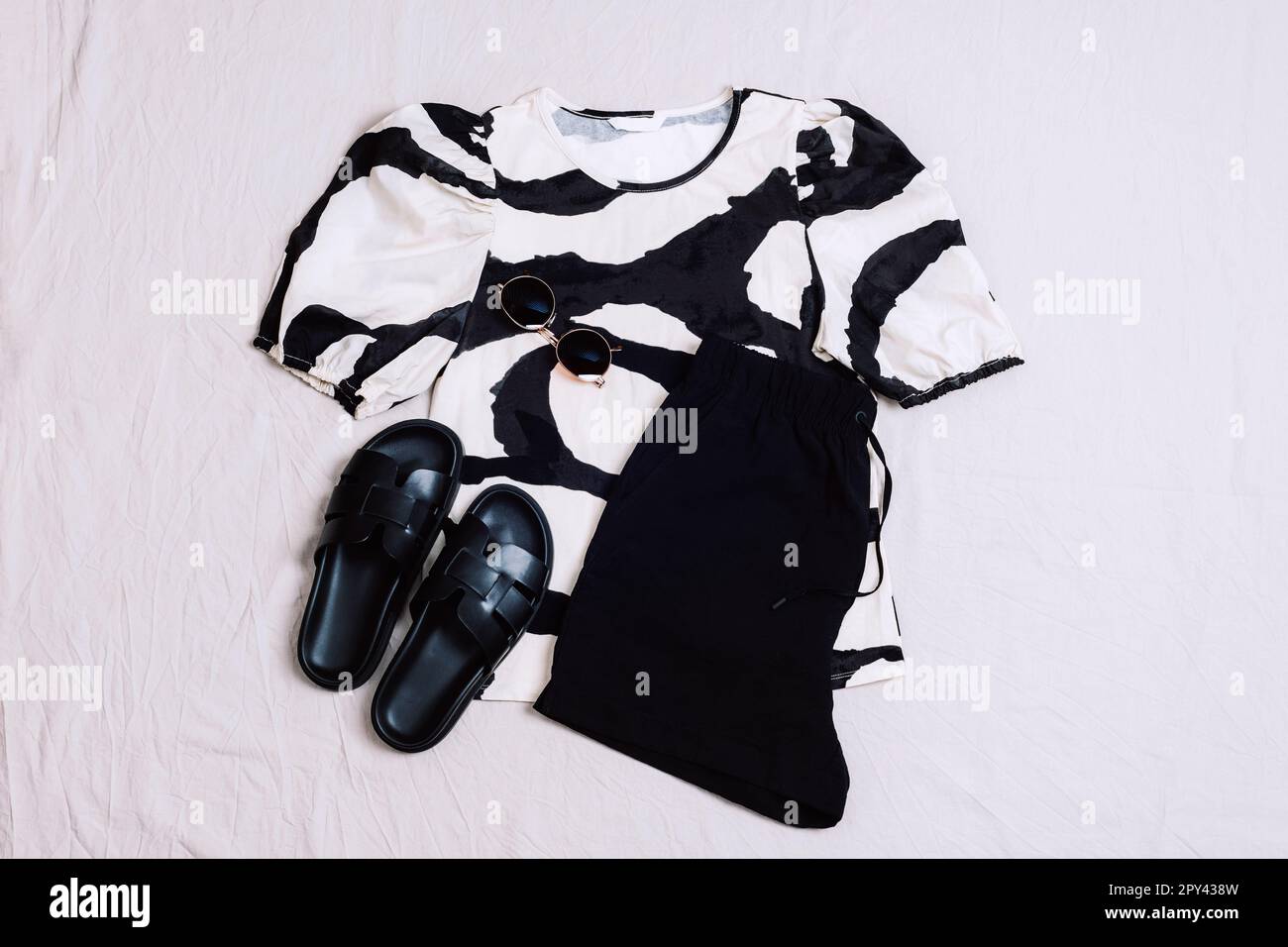 Schwarz-weiße Bluse, schwarze Shorts, schwarze Sandalen und Sonnenbrille auf grauem Hintergrund. Blick von oben auf die Freizeitkleidung der Frau. Trendiges, stilvolles Summ Stockfoto
