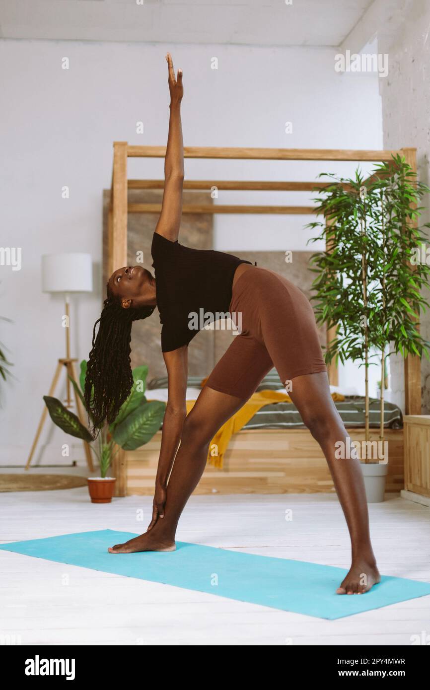 Fit Afro american Woman beugt Körper an Bein mit ausgestreckten Armen und macht Yoga auf einer Turnmatte im dekorierten Studio. Muskeldehnung, Gymnastik Stockfoto