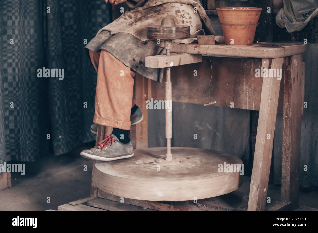 Handwerker in schmutzigen Arbeitskleidung, sitzt auf Töpfermaschine. Modellgeschirr auf Rädern in der Töpferwerkstatt. Stockfoto