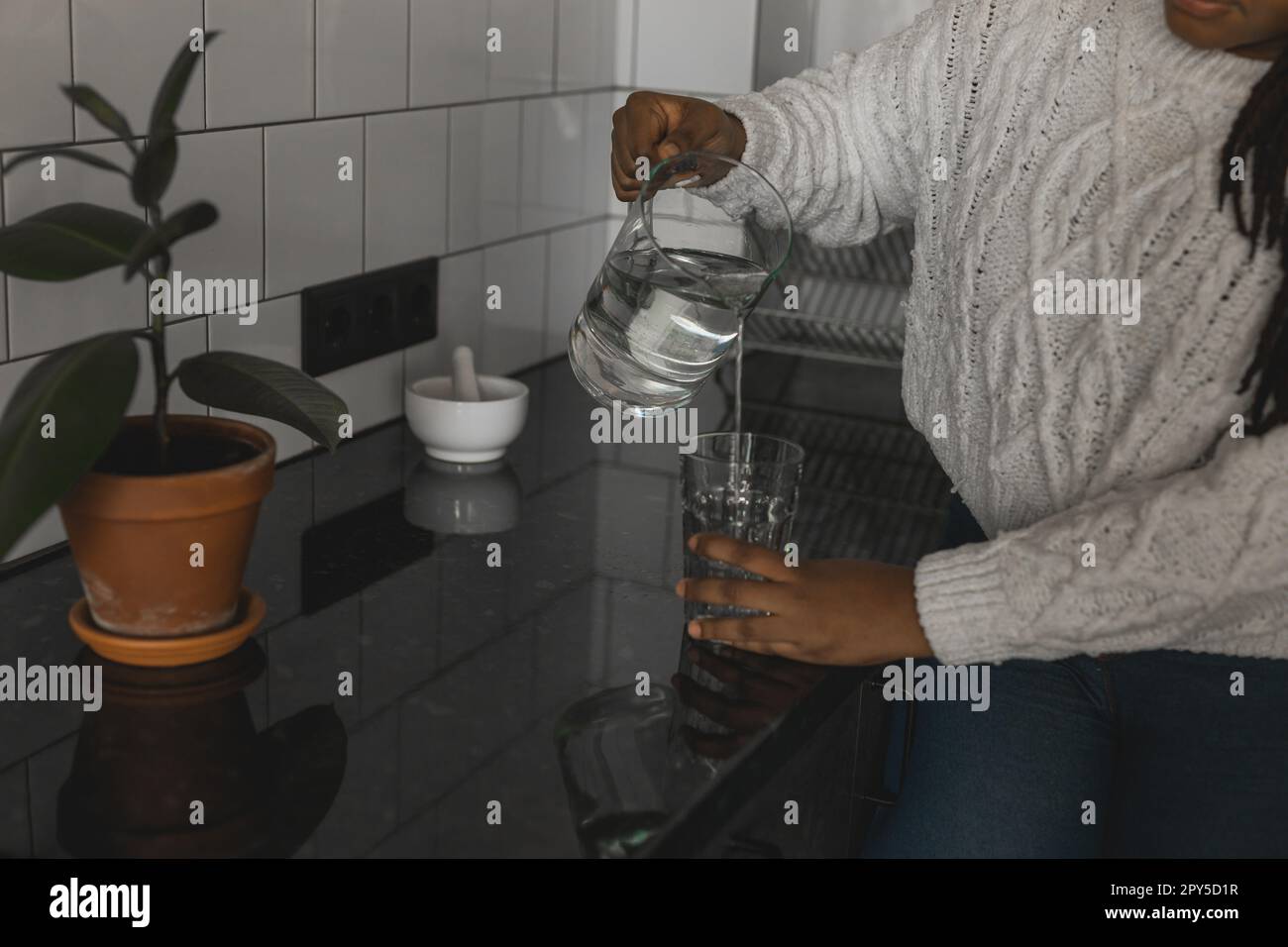 Aus nächster Nähe durstige afroamerikanische Frau, die Mineralwasser in der Küche trinkt, gesunde Flüssigkeit von Kanne zu Glas gießt, glückliche Frau mit Zöpfen, die erfrischende Getränke genießt Stockfoto