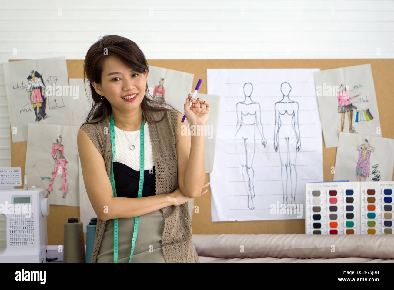 Junger Modedesigner, der einen magischen Stift mit einem Lächeln hält. Muster für den Katalog mit Tüchern und Farbproben befinden sich auf Korkplatten. Asiatische Frau, die in der Textilfabrik arbeitet. Stockfoto