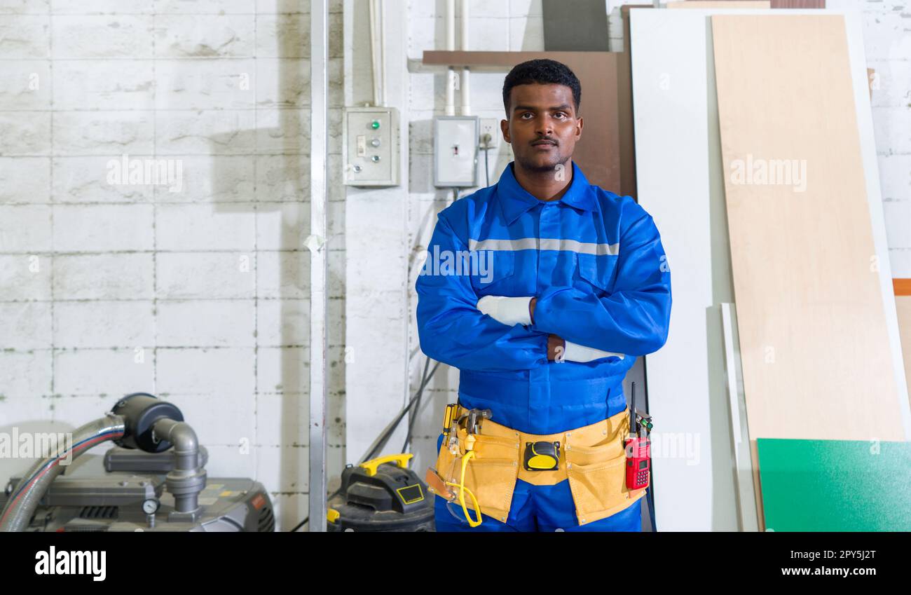 Junger Techniker in blauem Mechaniker-Overall, Schutzhandschuh und gelben Werkzeuggürteln stehen mit gekreuzten Armen in einer Möbelfabrik aus Holz. Stockfoto