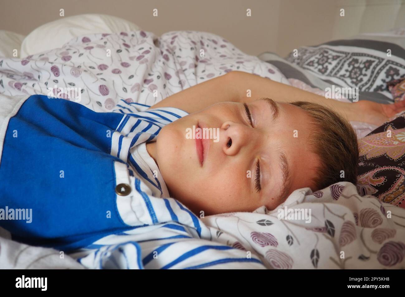 Ein wunderschöner, 10 Jahre alter, weißer Junge mit blondem Haar, gekleidet in gestreiften Pyjamas, schläft auf dem Bett mit einer flauschigen Decke, umarmt ein Kissen. Sanftes Licht der Morgensonne strömt durch das Fenster. Stockfoto