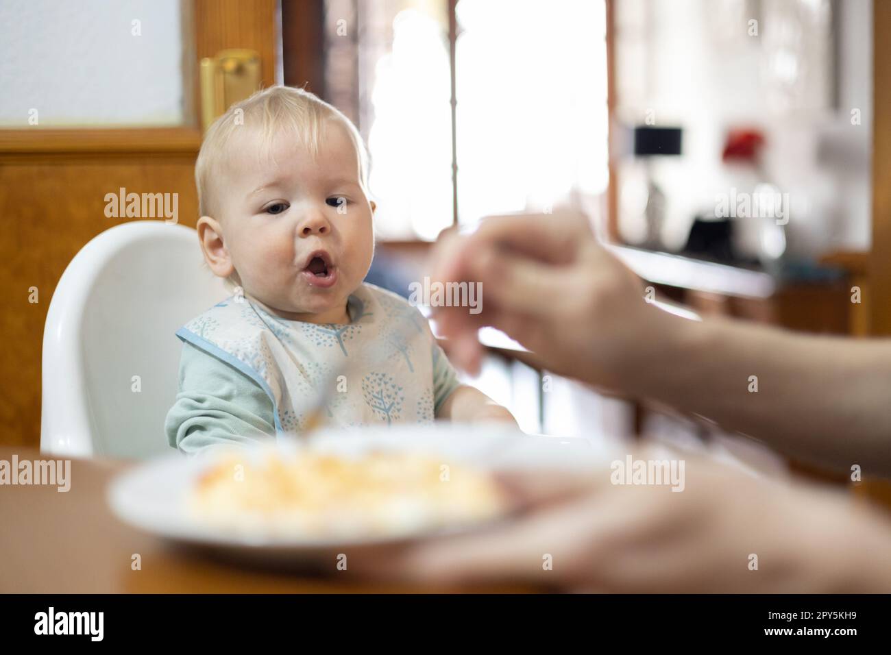 Mutterlöffel füttert ihr Baby Kind, das in einem Hochstuhl am Esstisch in der Küche zu Hause sitzt Stockfoto