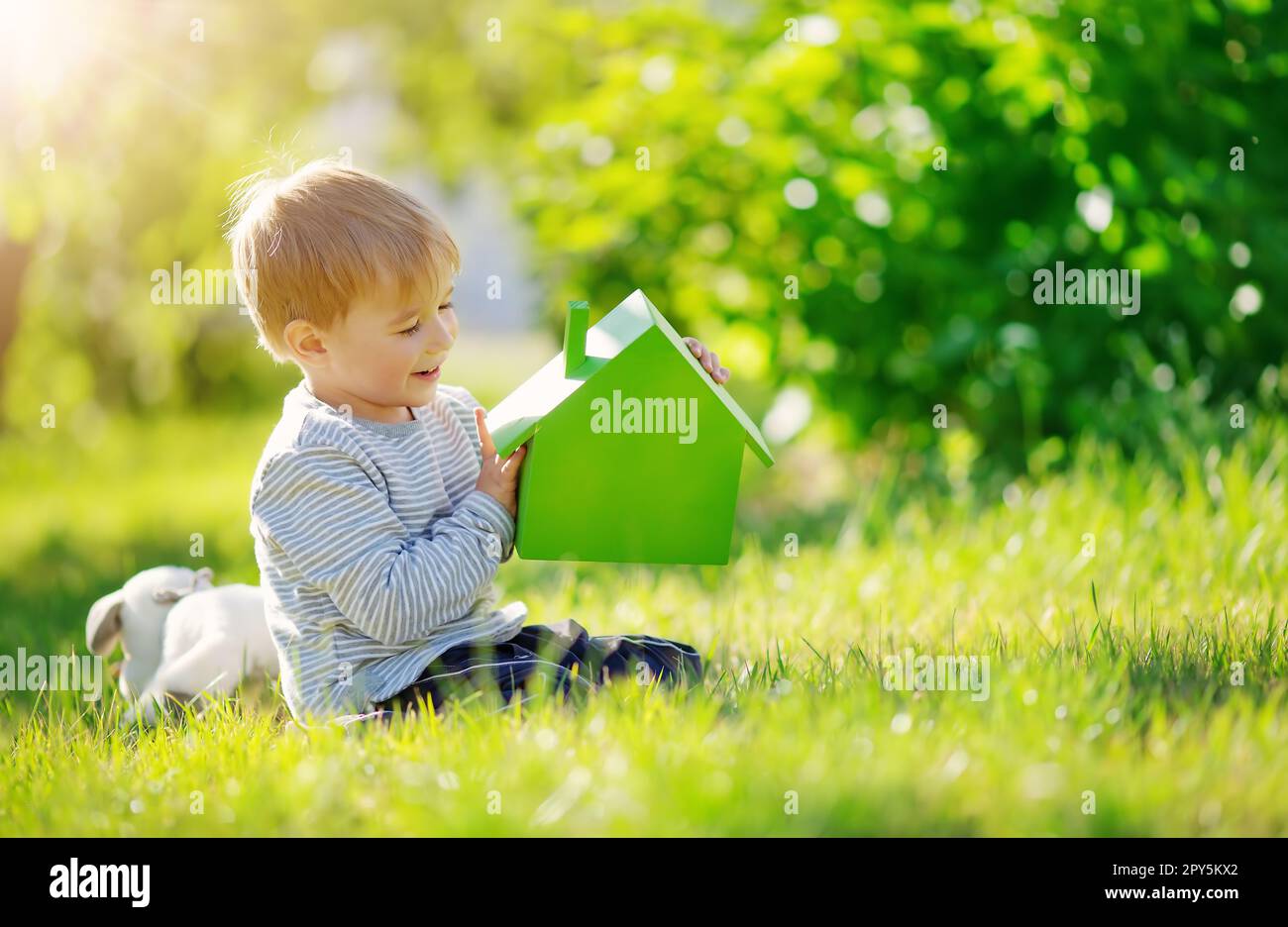 Ein lächelndes Kind, das auf der Frühlingswiese sitzt, mit einem Modell des grünen Hauses in seinen Händen. Stockfoto