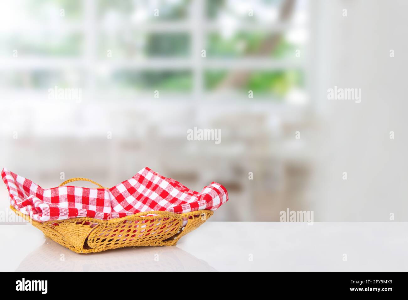 Leerer Picknickkorb. Nahaufnahme eines leeren Strohkorbs mit einer karierten roten Serviette auf dem Tisch über verschwommenem Küchenhintergrund. Für die Montage Ihrer Lebensmittel- und Produktpräsentation. Stockfoto