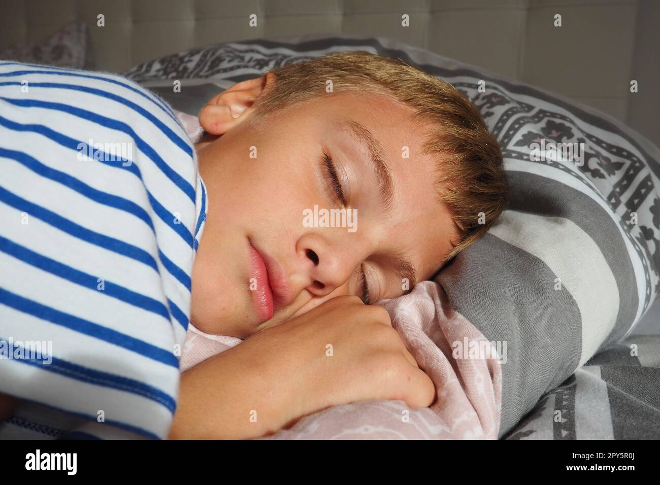 Ein wunderschöner, 10 Jahre alter, weißer Junge mit blondem Haar, gekleidet in gestreiften Pyjamas, schläft auf dem Bett mit einer flauschigen Decke, umarmt ein Kissen. Sanftes Licht der Morgensonne strömt durch das Fenster. Stockfoto