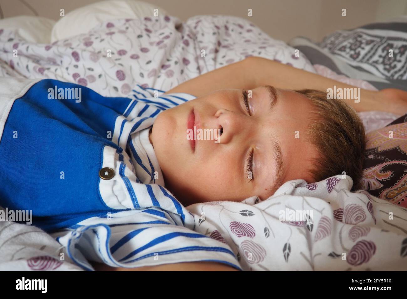 Ein wunderschöner, 10 Jahre alter, weißer Junge mit blondem Haar, gekleidet in gestreiften Pyjamas, schläft auf dem Bett mit einer flauschigen Decke, umarmt ein Kissen. Sanftes Licht der Morgensonne strömt durch das Fenster. Stockfoto