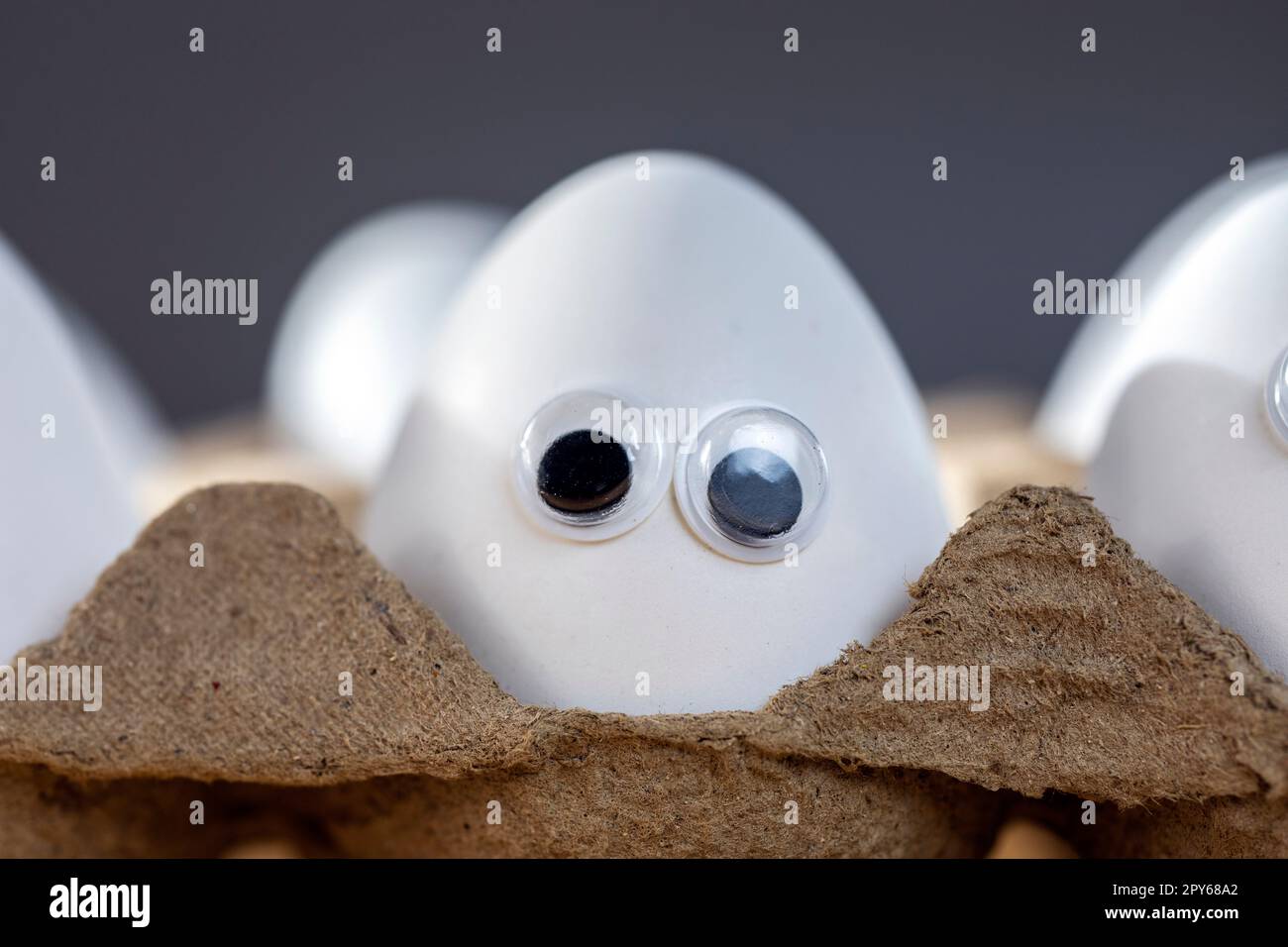 Lustige Gesichter auf weißen Eiern in einer Schachtel mit Bio-Hühnereiern auf dem Küchentisch, die große Animationsaugen zusammenschließen. Humor, Essen und osterferienkonzept. Stockfoto