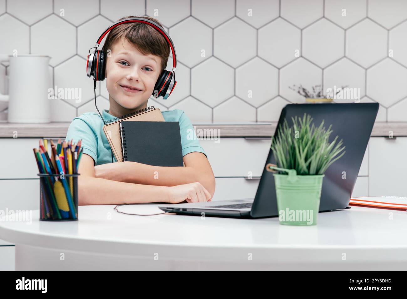 Porträt eines jungen, süßen Jungen, der T-Shirt, Kopfhörer trägt, vor dem Laptop sitzt, kuschelige Notizbücher in der Hand hält. Stockfoto