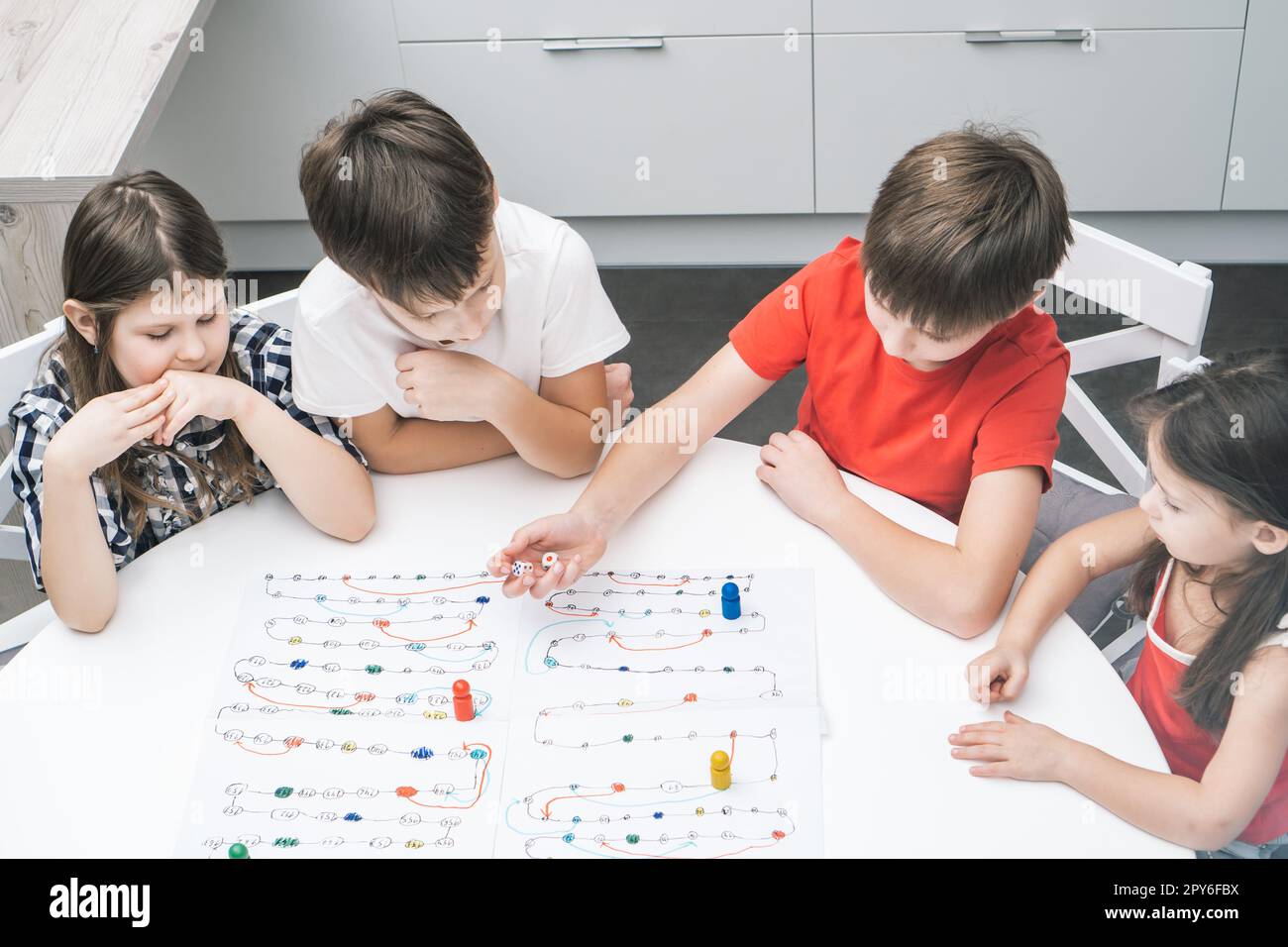 Freunde spielen Brettspiel mit Würfel und Chips auf dem Tisch, von oben gesehen. Kinder schauen sich den Jungen an, würfeln und bewegen die Figur. Stockfoto