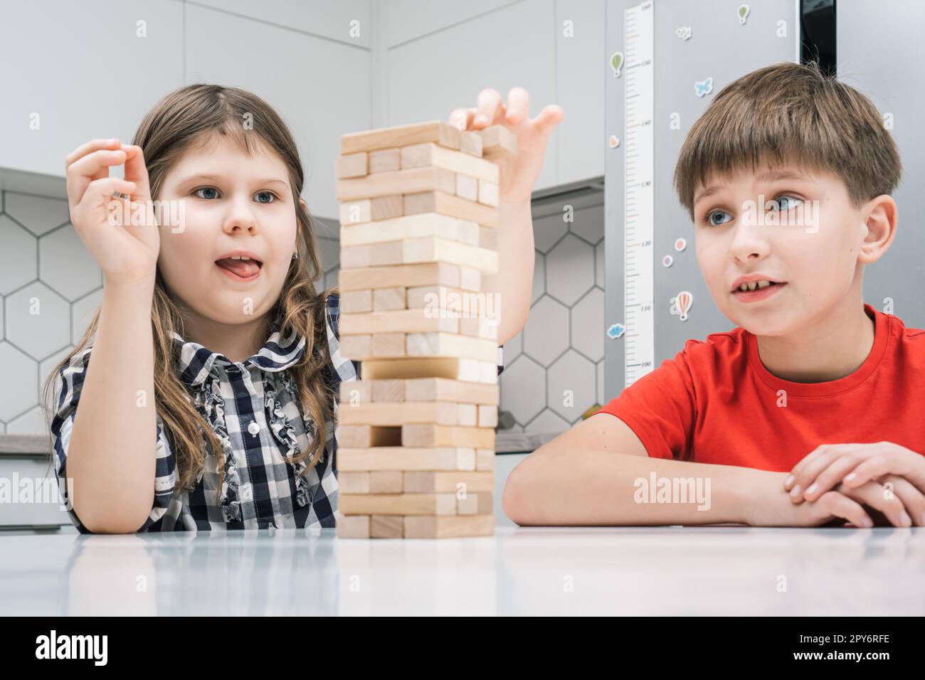 Kinder spielen Jenga, sitzen am Tisch. Junge und Mädchen mit konzentriertem Gesichtsausdruck bauen Turm aus Holzblöcken. Stockfoto