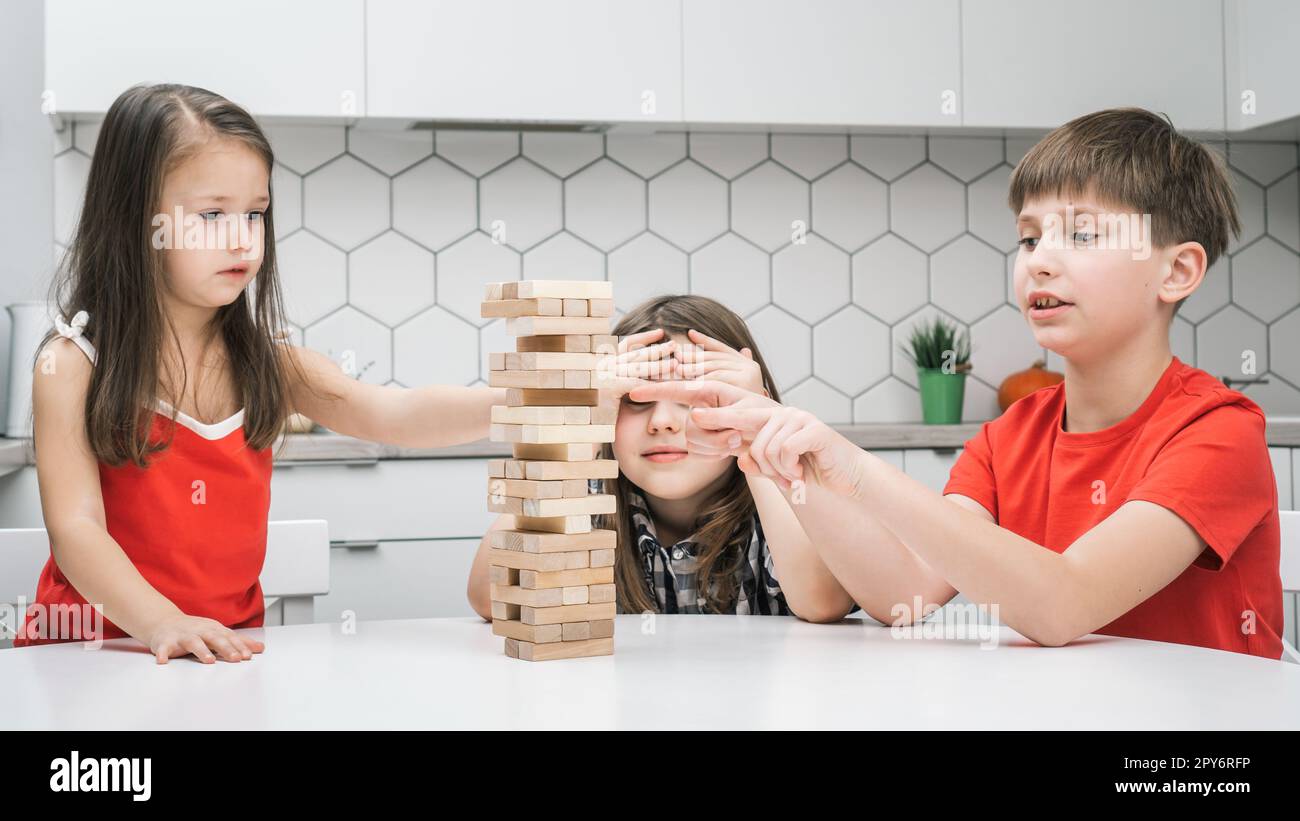 Glückliche Schulkinder spielen Turm, sitzen am Küchentisch. Süße Jungen und Mädchen bauen einen Turm aus kleinen Holzblöcken. Stockfoto