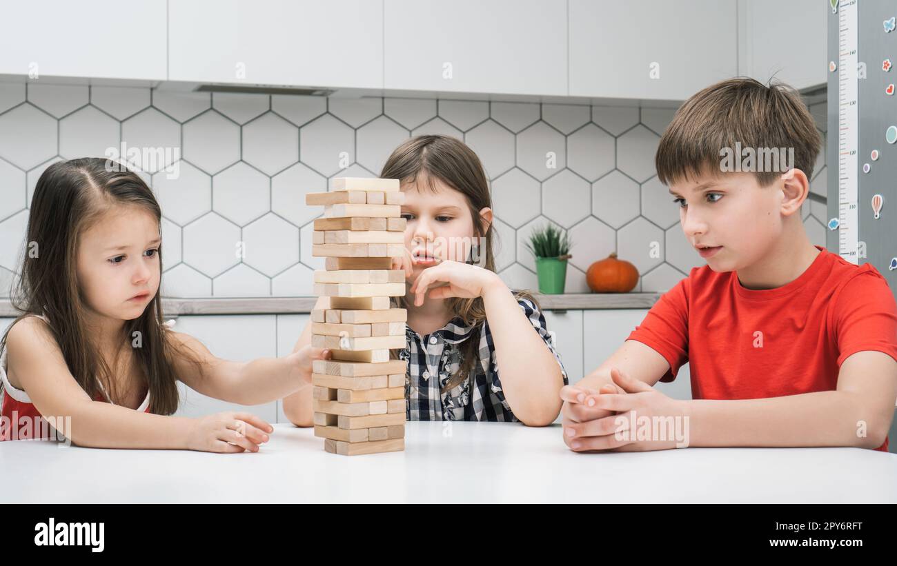 Konzentrierte Schulkinder spielen Turm, sitzen am Küchentisch. Junge und Mädchen bauen Turm aus kleinen Holzblöcken. Stockfoto