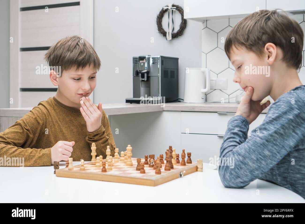 Denkt, Schuljungen spielen Schach, sitzen am Küchentisch, Seitenansicht. Holzschachbrett mit schwarz-weißen Figuren. Stockfoto