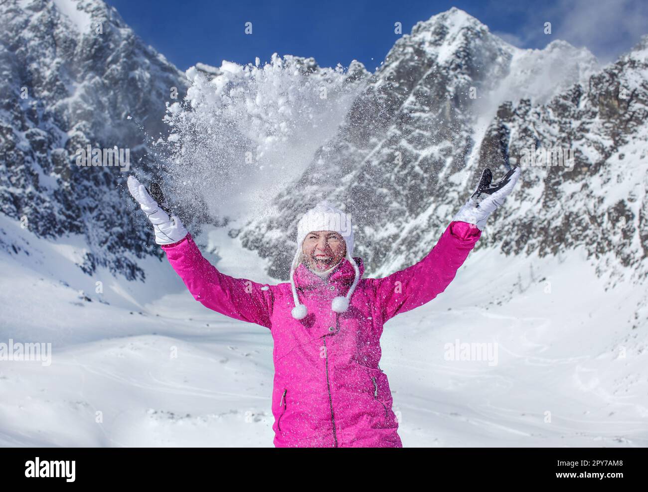 Junge Frau in pinkfarbener Skijacke, Handschuhen und Wintermütze, lächelt, wirft Schnee in die Luft, Sonne scheint auf den Berg hinter ihr. Skigebiet Skalnate Pleso, Slowakei. Stockfoto