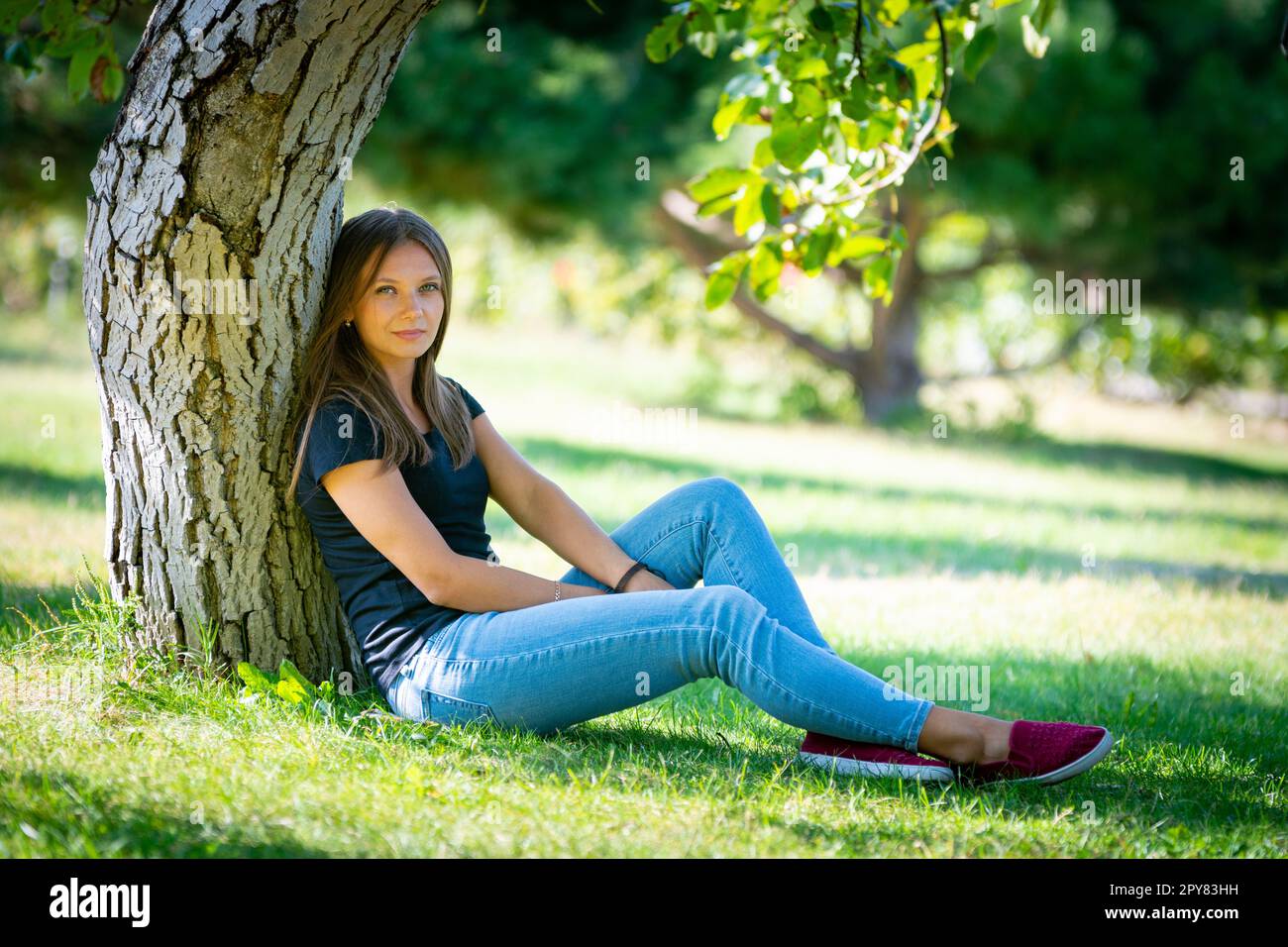 Ein Mädchen sitzt unter einem Baum in einem sonnigen Park und schaut in den Rahmen Stockfoto