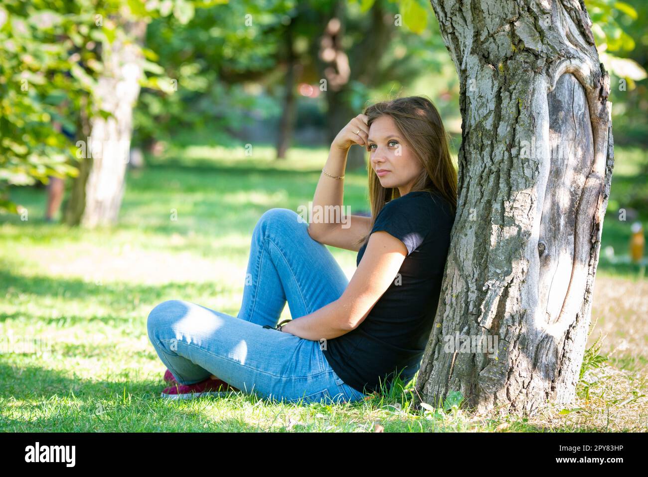 Ein Mädchen sitzt unter einem Baum in einem sonnigen Park und blickt gedankenvoll in die Ferne Stockfoto